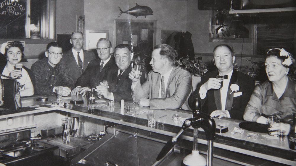 Eight customers along the front bar raising their glasses, with a mounted tuna on the wall behind